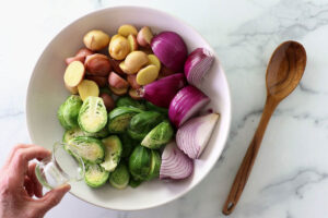 Hand pours oil into bowl of potatoes, onions, and Brussels sprouts on marble.