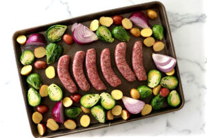 Sheet pan with uncooked sausages, Brussels sprouts, red onion, and potatoes.