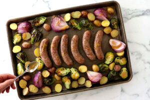 Tray with sausages, Brussels sprouts, onions, potatoes; hand drizzles oil over vegetables.