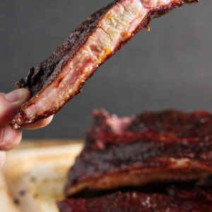 Hand holding saucy smoked rib, more ribs and asparagus on wooden board.