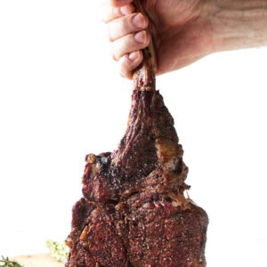 Hand holding cooked tomahawk steak over rosemary-garnished wooden board.
