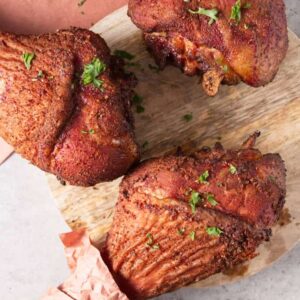 Three smoked turkey legs with parsley on a wooden board, one wrapped.