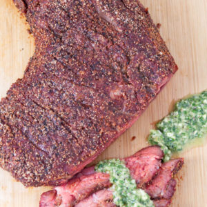 Sliced smoked tri-tip with peppery crust and chimichurri on wooden board.