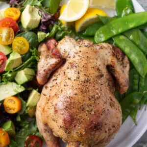 Smoked game hen on a plate with greens, tomatoes, avocado, and lemon wedges.