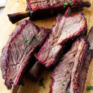 Four smoked beef ribs with dark crust on a wooden cutting board.