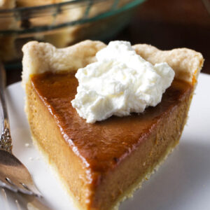 Pumpkin pie slice with whipped cream on plate, fork beside it.