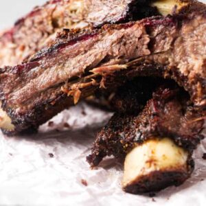 Two smoked beef ribs with dark crust on parchment, juicy meat visible.