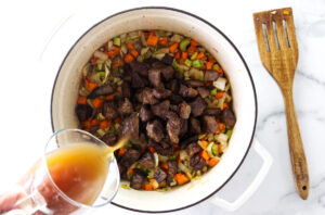 Pouring broth into a pot with chunks of beef and veggies.