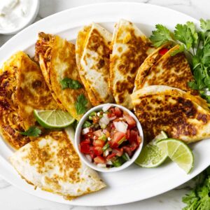 Several sliced quesadillas on a platter with pico de gallo and limes.
