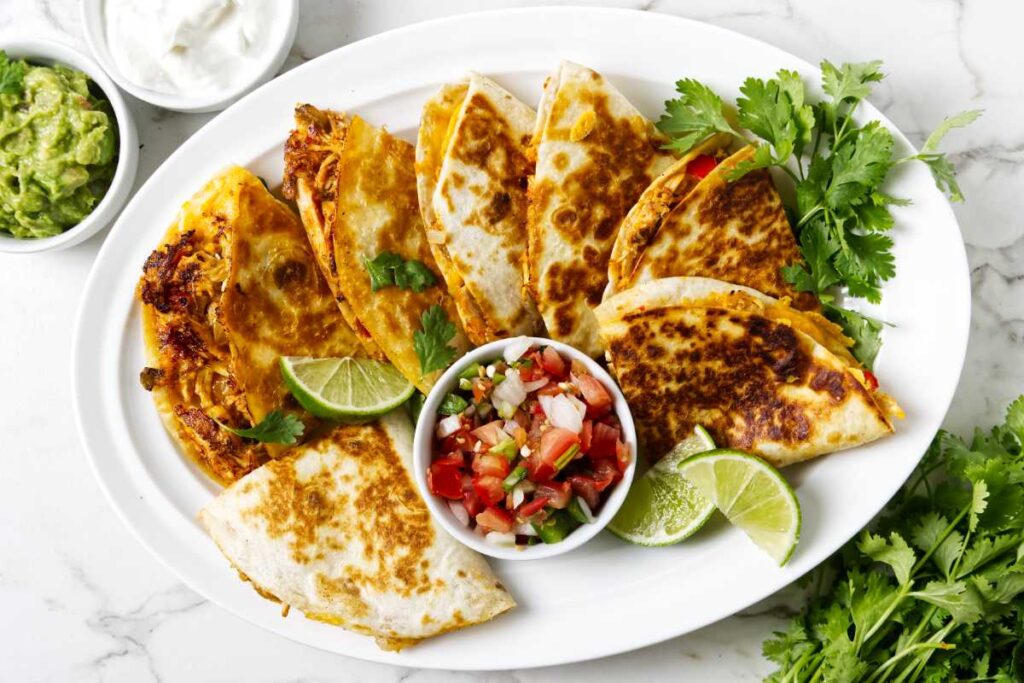 Quesadilla slices with salsa, lime, cilantro; guacamole and sour cream on marble.