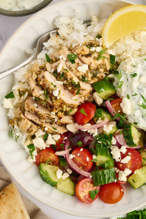 Bowl of Greek chicken with rice, cucumber tomato salad, feta, and herbs.