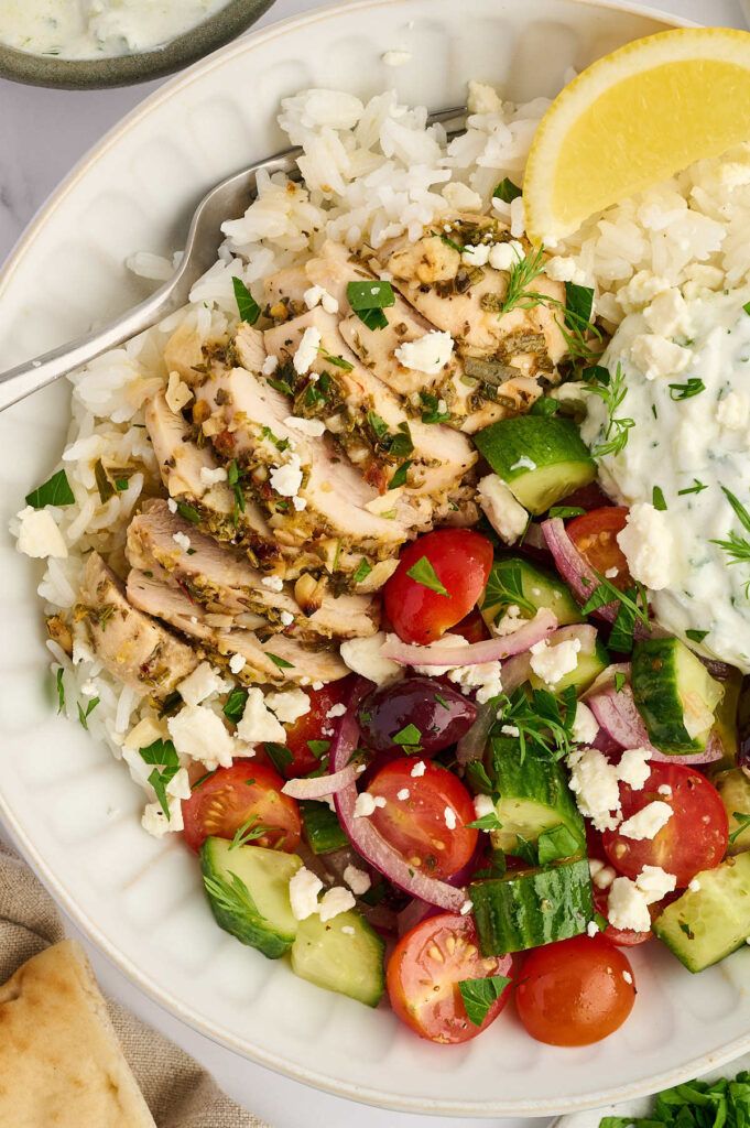 Bowl of Greek chicken with rice, cucumber tomato salad, feta, and herbs.
