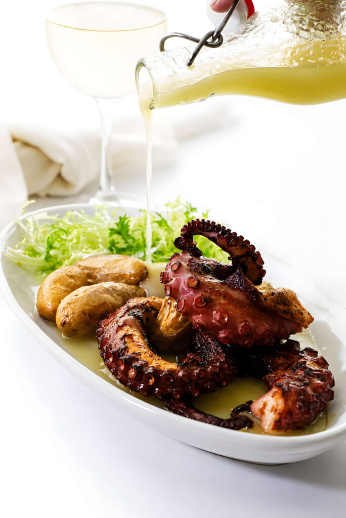 Grilled octopus and potatoes garnished with greens, sauce poured, wine glass nearby.