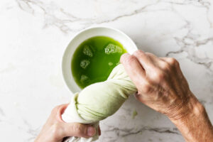 Hands squeezing green liquid from cloth into bowl on marble counter.
