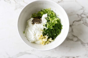 White bowl of yogurt with cucumber, garlic, dill, and pepper on marble.