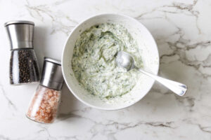 Bowl of tzatziki dip with spoon beside pepper and salt grinders.