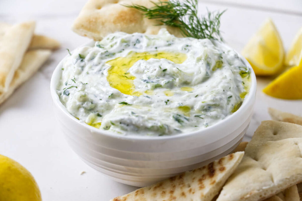 Bowl of tzatziki with olive oil, dill, pita, and lemon wedges.