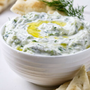 Bowl of tzatziki with olive oil, dill, and pita bread around.
