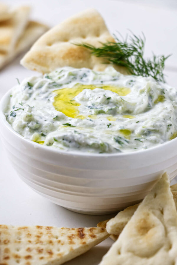 Bowl of tzatziki with olive oil, dill, and pita bread around.