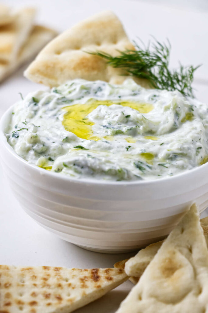 Bowl of tzatziki with olive oil, dill, and pita bread around.