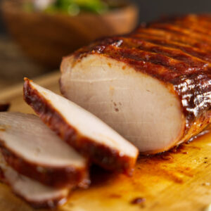 Sliced glazed pork roast on cutting board, vegetables blurred behind.