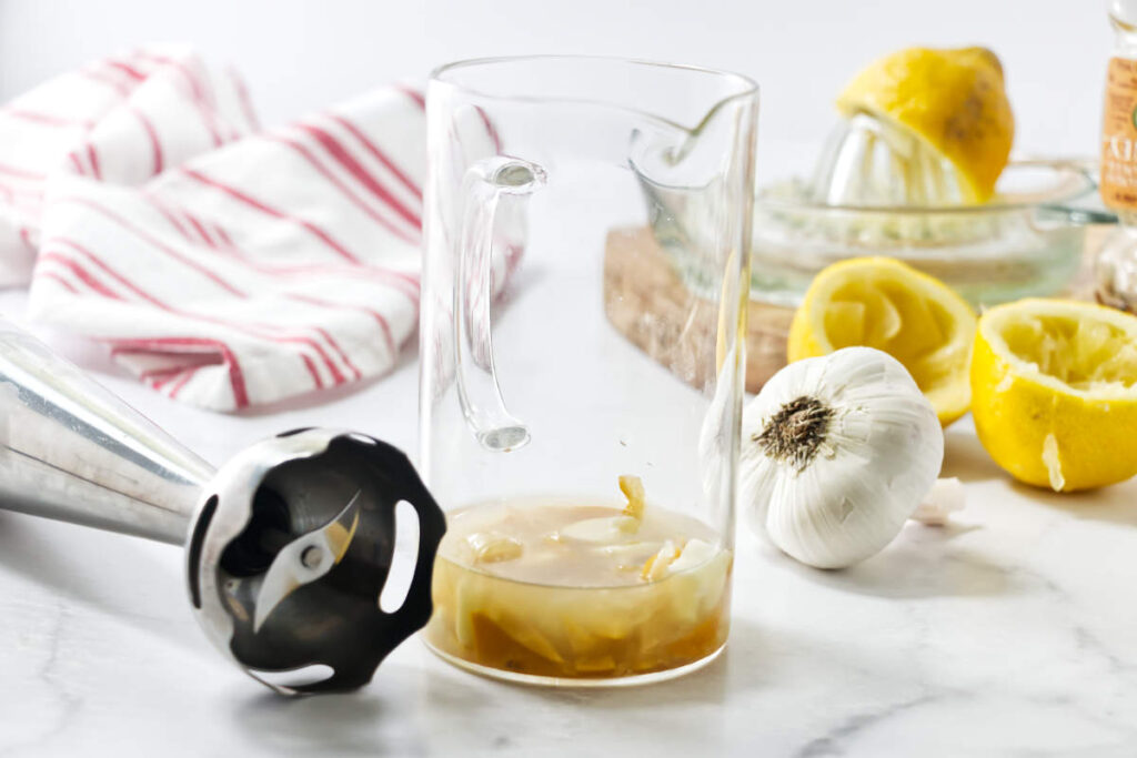 Blender stick and measuring cup with garlic, lemon, and liquid on countertop.