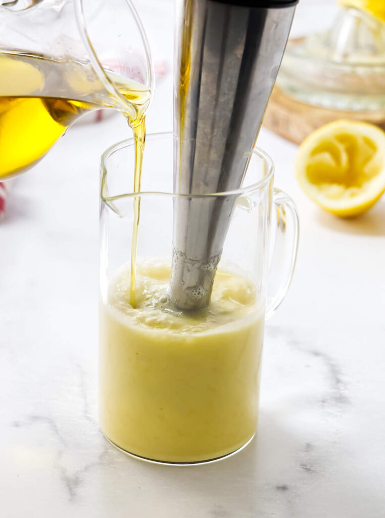 Drizzling olive oil into a jar while an immersion blender blends preserved lemons and lemon juice.
