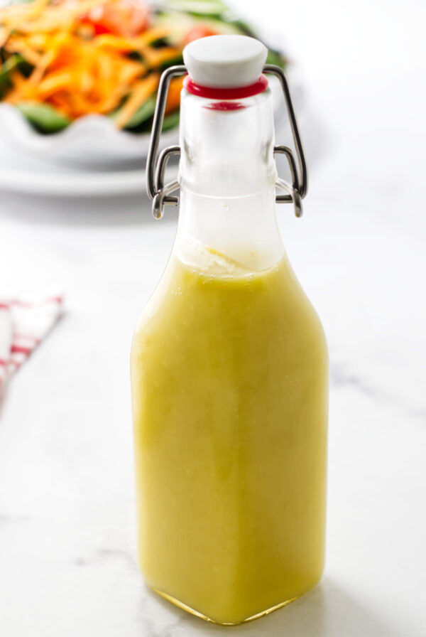 Glass bottle of preserved lemon salad dressing on white surface, salad blurred behind.