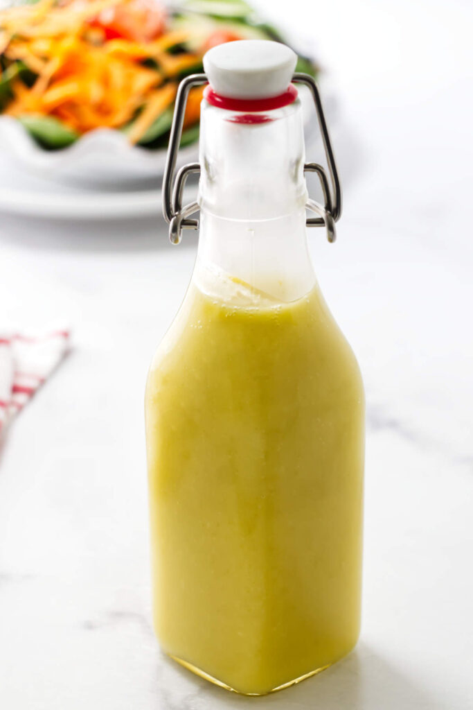 Glass bottle of preserved lemon salad dressing on white surface, salad blurred behind.