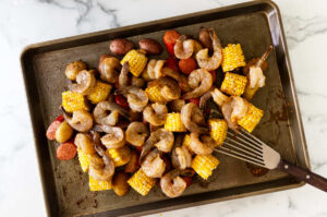 Broiled shrimp turning pink on top of roasted potatoes, corn, and sausage.