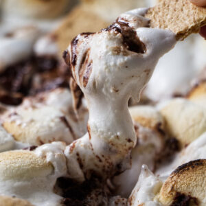 Hand dipping graham cracker into gooey s&rsquo;mores dip, close-up view.