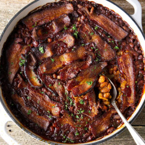 White pan of baked beans with bacon and herbs on rustic wood.
