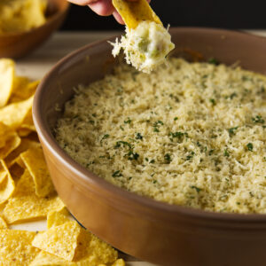 Hand dipping tortilla chip into cheesy dip with chips around dish.