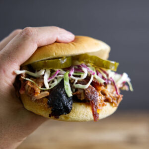 Hand holds pulled pork sandwich with slaw and pickles; blurred background.