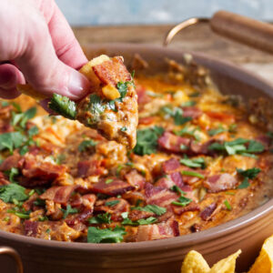 Hand dipping tortilla chip into cheesy bacon dip with herbs.