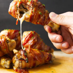 Hand holding a cheesy bacon-wrapped drumstick, others on wooden board.