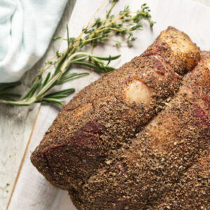 Seasoned prime rib on white cutting board with rosemary and thyme.