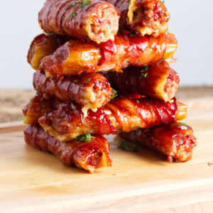 Bacon-wrapped cheese sticks glazed with sauce on a wooden cutting board.