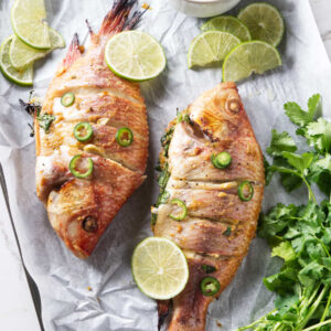 Two grilled tilapia with chili, lime, cilantro, and sauce on parchment.