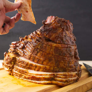 Hand lifting crispy skin from spiral-cut baked ham on cutting board.