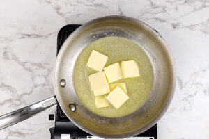 Stainless steel pan melting butter cubes on stovetop over marble counter.