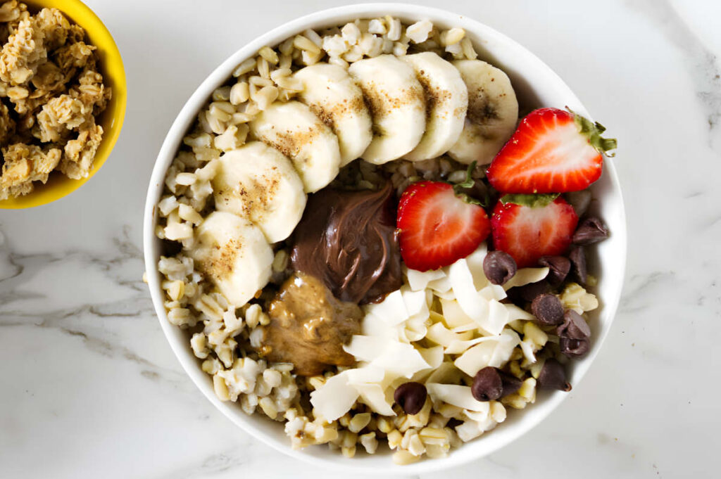 Whole oats in a bowl with fruit, chocolate toppings, and granola on marble surface.