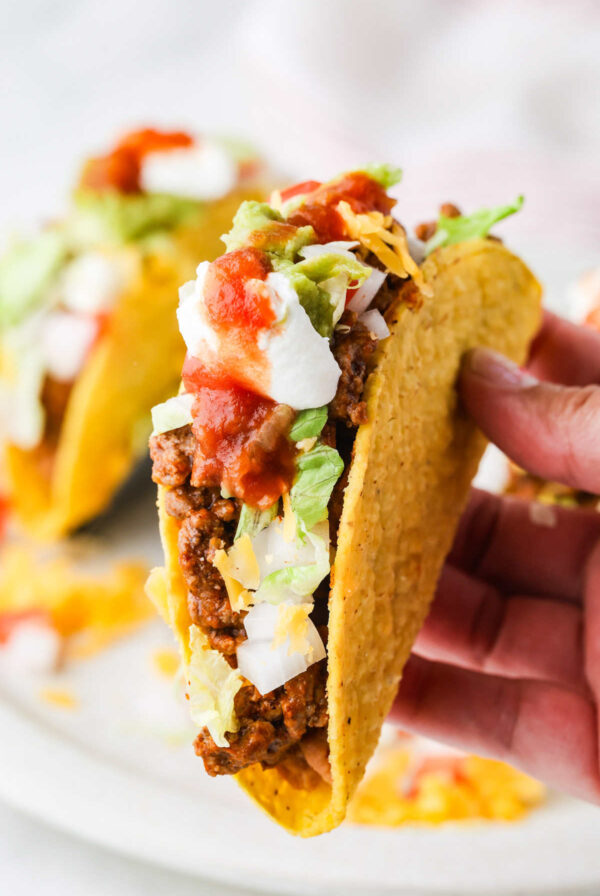 Hand holding a crisp taco filled with seasoned ground beef and toppings.