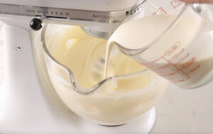 Measuring cup pours cream into white stand mixer with creamy batter.