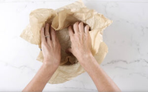 Hands pressing brown parchment paper into a bowl on marble surface.