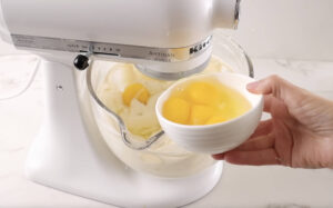 Hand holds bowl of eggs beside white stand mixer with batter.