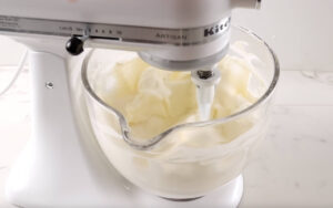 Stand mixer whipping cream in glass bowl on white counter.