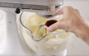 Hand pours vanilla extract into stand mixer with creamy batter.