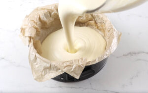 Cake batter being poured into a parchment-lined round pan on marble.