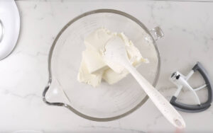 Mixing bowl with cream cheese, spatula, and mixer paddle on countertop.